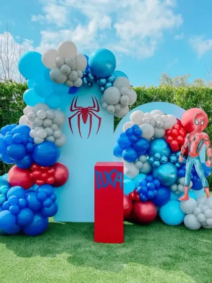 An outdoor Spider-Man themed birthday party. The setup uses two blue backdrops with the Spider-Man logo and a red, blue, and grey balloon arrangement to create a superhero scene.