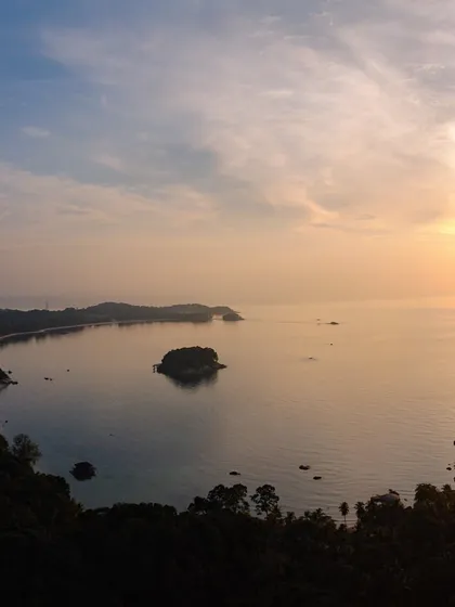 A panoramic drone shot of a serene bay on Bintan Island, Indonesia, at sunset, with a small island in the middle of the calm water.
