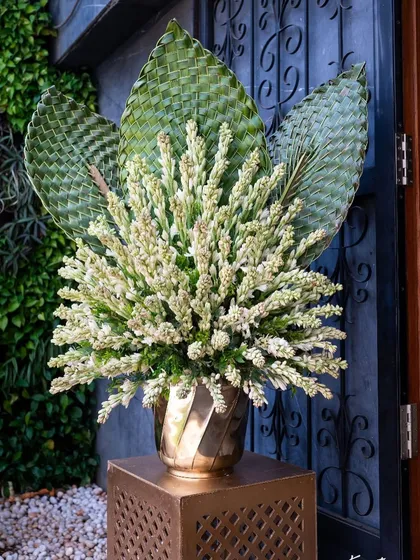A floral arrangement featuring white tuberoses and woven palm leaves in a brass vase, a detail from a traditional wedding.