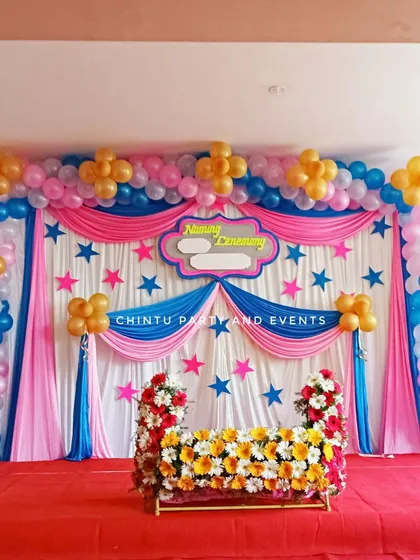 A complete stage setup for a naming ceremony with a decorated cradle. The backdrop features pink and blue drapes under a full balloon arch.