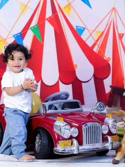 This little boy is standing proudly next to a red toy car in our fun carnival setting, ready for his birthday adventure.