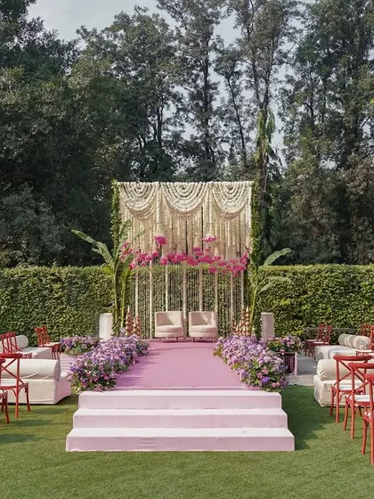 The mandap for a garden wedding, featuring a backdrop of traditional Indian 'jaal' work and a cascade of flowers. The mix of red chairs and pastel decor creates a unique, eclectic look.