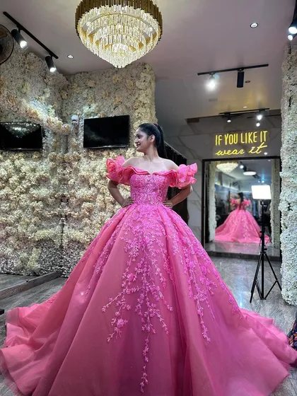 Step into my Delhi studio and find your dream dress. This vibrant fuchsia pink ball gown, with its off-shoulder ruffled sleeves and intricate floral embroidery, is a perfect example of the show-stopping pieces you can try on. The studio is designed to be a magical space for your fitting experience.