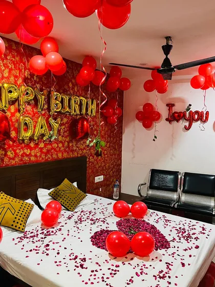 A birthday decoration in a hotel room, featuring gold 'Happy Birthday' foil balloons against a vibrant red patterned wallpaper, with red balloons scattered on the bed.