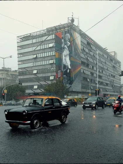 A Kaali-Peeli taxi navigates a wet intersection, with a large, modern building featuring a mural in the background. This image captures the mix of old and new Mumbai on a rainy day.