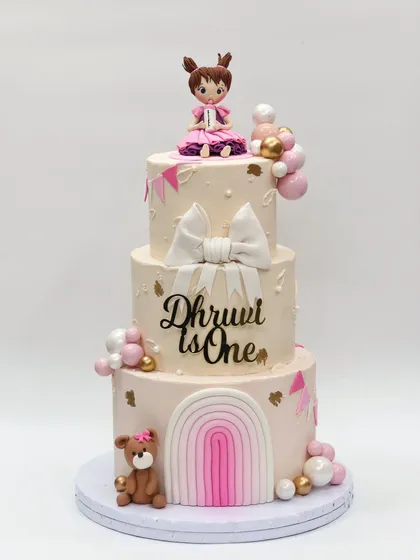 A grand three-tier first birthday cake with a soft pink and white theme, featuring a large bow, a rainbow, a teddy bear, and a girl figurine.