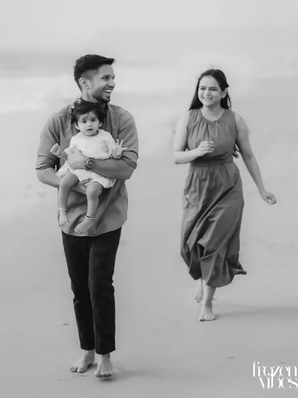 A black and white photograph of a family walking along the shore. The father carries their child while the mother walks alongside, creating a timeless image of a family on the move.