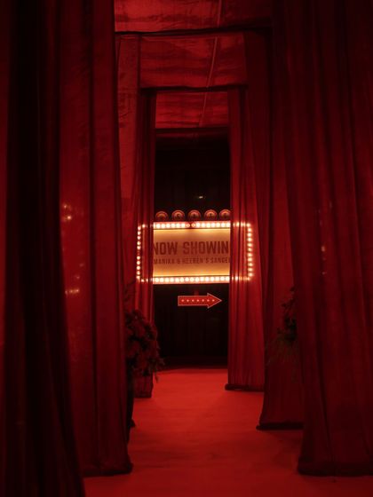 The dramatic entrance walkway for the Sangeet, lined with heavy red velvet curtains, leading guests towards the glowing marquee sign.