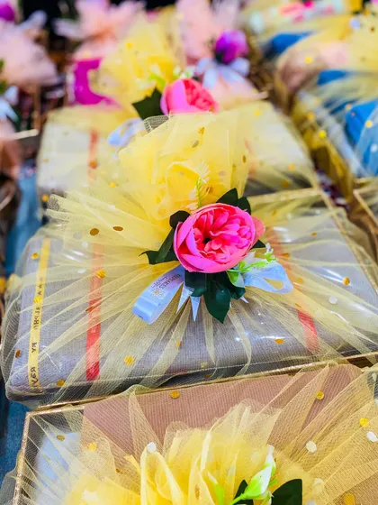 A collection of wedding favours wrapped in bright yellow tulle. Each package is adorned with a vibrant pink peony, making for a cheerful and eye-catching gift for guests at a mehandi or haldi ceremony.