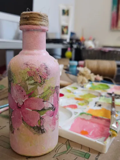 A finished decoupage bottle decorated with soft pink textures and floral designs, sitting on my messy-but-loved workspace, demonstrating my custom decor services.