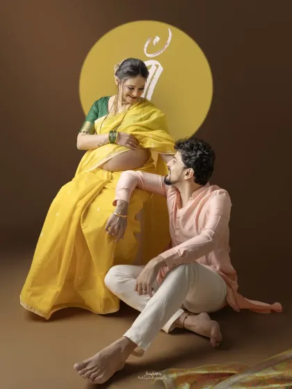The couple shares a loving look during their traditional maternity session. The vibrant yellow saree and the 'Maa' projection create a visually stunning and meaningful composition.