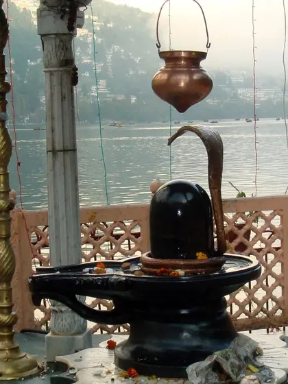 A Shiva Lingam installed by a tranquil lake, merging the elements of water and earth in worship. I guide you on how to connect with the five elements through devotion to Lord Shiva.