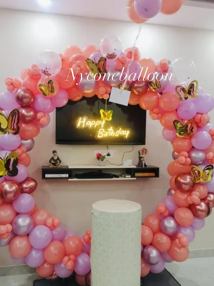 This beautiful butterfly-themed ring decoration is perfect for birthdays or anniversaries. The arrangement uses peach, pink, and lavender balloons with decorative butterfly accents and a central 'Happy Birthday' neon sign.