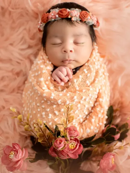 A newborn baby wrapped in a peach-colored swaddle, lying on a soft pink fur blanket and adorned with a floral headband.