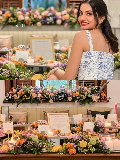 A vibrant grazing table overflowing with colorful flowers. This setup for an intimate gathering shows a woman smiling, surrounded by a beautiful arrangement of florals and personalized details.