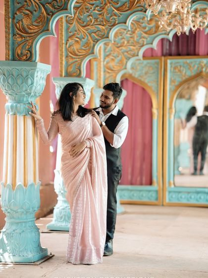A romantic pose against a grand, palace-like set with ornate blue pillars, the couple dressed in elegant traditional Indian wear.