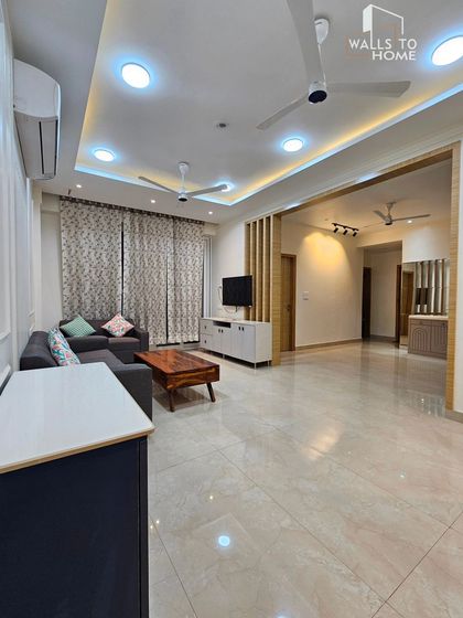 A wide view of a minimalist living room I designed at Supertech Emerald Court. The space features a comfortable sofa, a simple TV unit with a wooden partition, and a clean false ceiling design with multiple light sources.