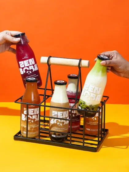 Two hands reach for bottles of Zen Boba from a stylish metal and wood carrier. This lifestyle shot against a bright, two-toned background conveys a sense of sharing and modern convenience.
