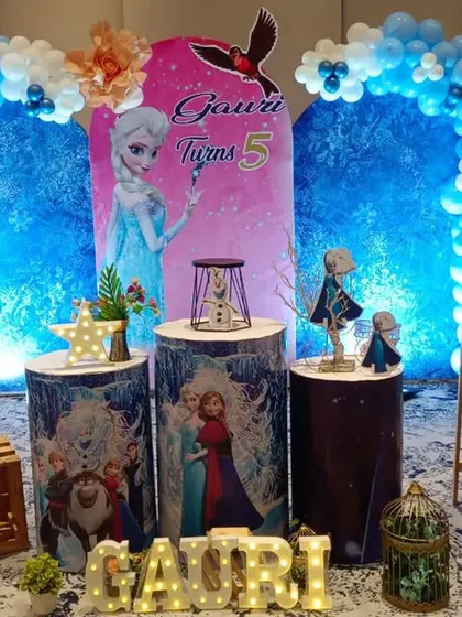 A Disney Frozen themed party is always a favorite. This setup for a fifth birthday includes a large blue balloon arch, cutouts of Elsa and Anna, and custom-printed display stands to create a complete winter wonderland scene.