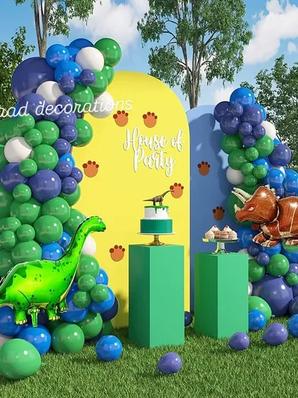 This outdoor dinosaur-themed party setup uses bold colors to create a prehistoric vibe. The scene is set with large dinosaur foil balloons, green pedestals for the cake, and two colorful balloon arches against matching backdrops.
