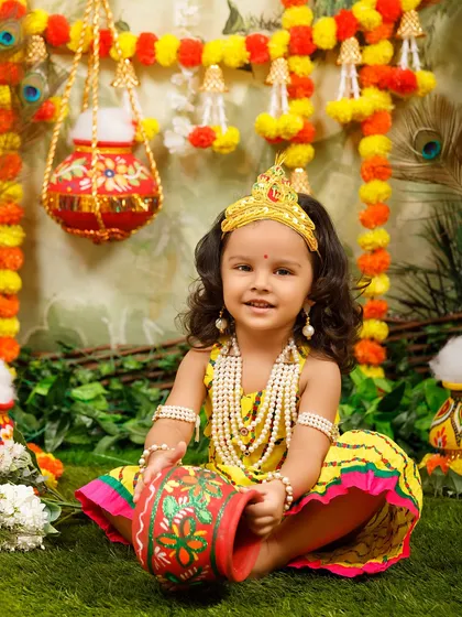 A sweet little Radha with curly hair and a crown, posing with her makhan pot.