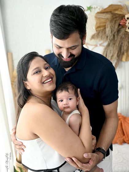 A candid and joyful family moment. The mother holds her newborn while the father embraces them both, all sharing a happy, natural smile in a bright, boho-inspired setting.