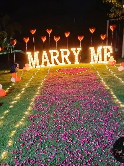 A grand outdoor proposal setup on a lush green lawn. The scene is lit by large, illuminated "MARRY ME" marquee letters, a pathway of scattered rose petals framed by fairy lights, and heart-shaped balloons.