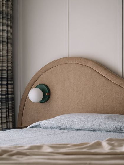 A close-up of the guest bed, showing the custom-designed globe sconce. The mix of textures, from the upholstered headboard to the plaid curtains, creates a cozy and inviting feel.