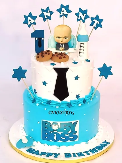 A sharp two-tier Boss Baby cake for a first birthday. The design features a fondant tie, cookies, a milk bottle, and star toppers with the baby's name.