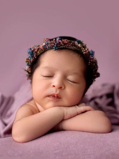 A beautiful portrait of a baby girl resting her head on her arms. The colorful headband adds a lovely detail against the simple purple backdrop.