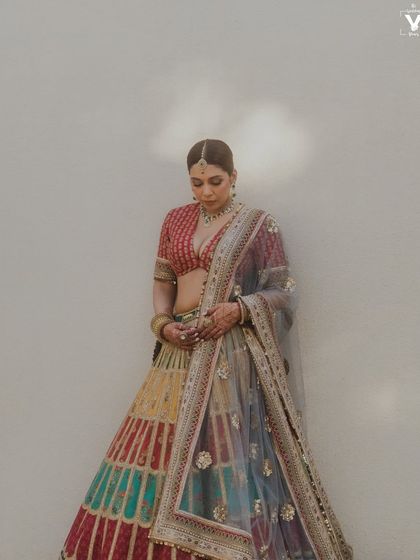 A full-length portrait of the bride in her multi-colored Sabyasachi lehenga. The soft light and simple wall background highlight the incredible detail and artistry of her outfit.