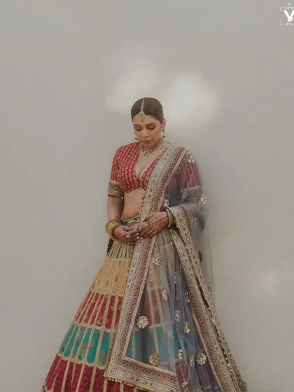 A full-length portrait of the bride in her multi-colored Sabyasachi lehenga. The soft light and simple wall background highlight the incredible detail and artistry of her outfit.