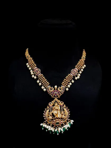 This is a classic temple jewellery long necklace, featuring a large Lakshmi pendant and intricate detailing. It is crafted with an antique gold finish and is perfect for wearing with silk sarees.