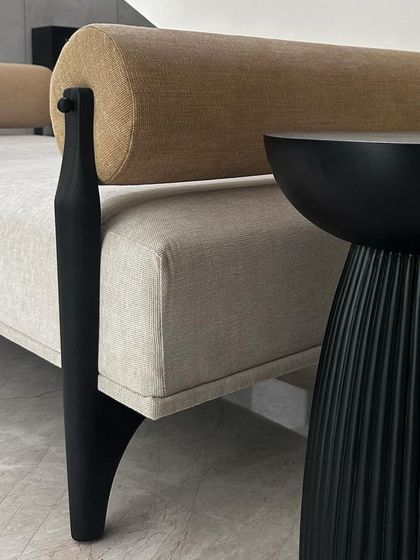 A detail shot of the day bench, showing the contrast between the smooth, dark-stained teak leg and the textured semi-suede upholstery of the main body and bolster pillow.