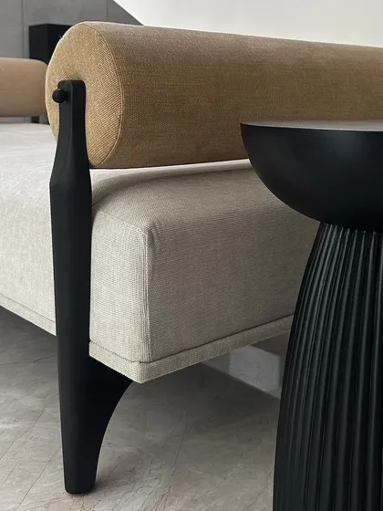 A detail shot of the day bench, showing the contrast between the smooth, dark-stained teak leg and the textured semi-suede upholstery of the main body and bolster pillow.