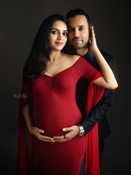 An elegant couple's maternity portrait against a dark grey backdrop. The woman's vibrant red gown and the intimate pose create a dramatic and romantic fine-art photograph.