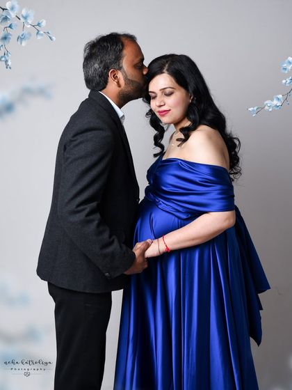 This portrait combines a vibrant blue satin gown with delicate floral elements in the background for a soft, romantic feel. The gentle kiss on the forehead adds a touch of tenderness to this elegant maternity photo.