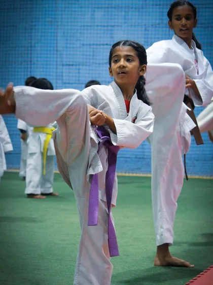 A female student with a purple belt practices a high kick, showing the level of skill and flexibility developed through our training programs.