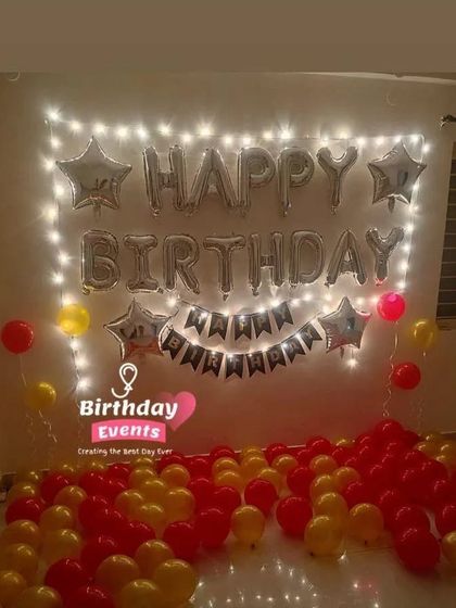 A simple and cheerful room setup with red and yellow balloons covering the floor, and a "Happy Birthday" banner framed by fairy lights.