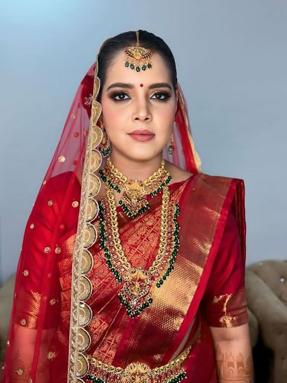 A classic South Indian bridal look. The makeup features a sharp, defined eye, a traditional bindi, and a bold lip, all complementing her red silk saree.