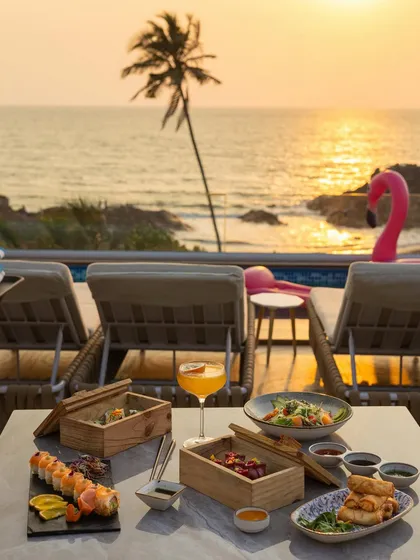 A stunning wide shot of a full meal set up against a golden sunset over the ocean. This is the ultimate lifestyle shot, selling a dream of luxury, travel, and exquisite dining.