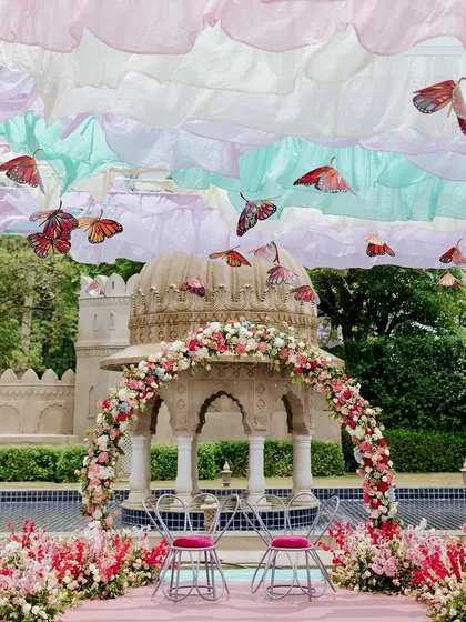 A magical, spring-inspired Mehendi theme. The couple's seating is set before a floral arch, with a dreamy sky of pastel ruffled drapes and fluttering butterflies creating a truly whimsical atmosphere.