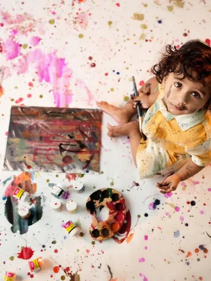 A playful and messy 'little artist' session, captured from above, showing a toddler sitting amidst colorful paint splatters with a palette and brushes.