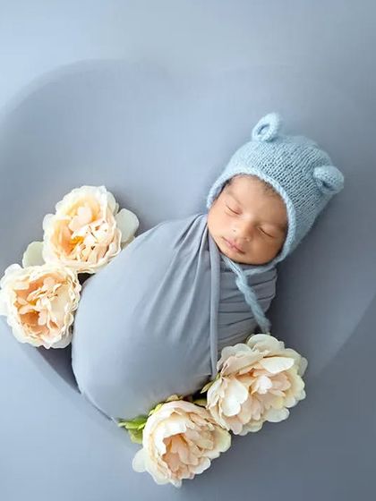 A sleeping newborn wearing a blue bear-ear bonnet is swaddled and placed in a grey heart bowl with delicate white flowers.