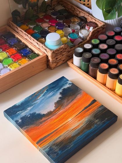 My finished sunset painting on a stretched canvas, placed on my desk next to my collection of gouache and acrylic paints. The bright orange and dark blue create a beautiful contrast.