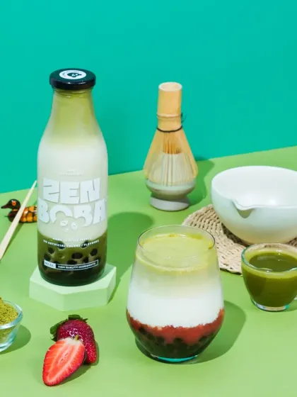 A bright and fresh composition for Zen Boba's matcha strawberry drink. The vibrant green background and neatly arranged ingredients like fresh strawberries and a matcha whisk communicate the product's natural and crafted quality.