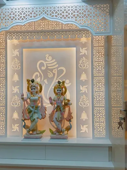 A pristine white Corian mandir featuring intricate jali work on the sides and top arch. The back panel is subtly engraved with religious symbols and a large Ganesha outline, all softly illuminated to create a tranquil and sacred space.