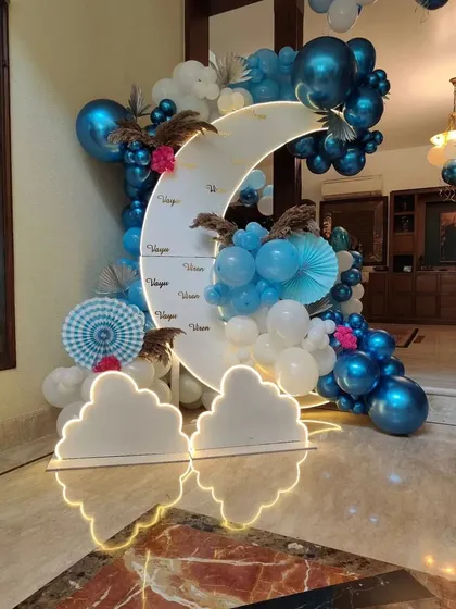 A dreamy moon-and-clouds theme for a welcome home celebration. This setup includes a large crescent moon prop, cloud cutouts with neon edging, and a blue and white balloon arrangement.