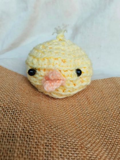 A close-up product shot of the chick amigurumi on a textured burlap surface. This shows the details of its little beak and eyes.