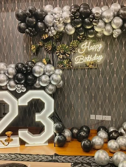 A chic and modern 23rd birthday decoration. The setup features a square balloon arch in black and silver, a "Happy Birthday" neon sign, and large light-up numbers.